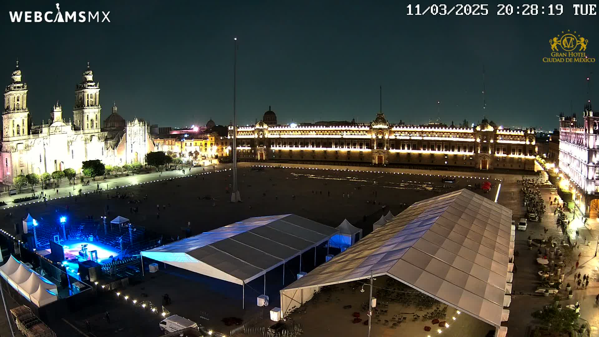 A nighttime, aerial view of a large plaza with a cathedral and government buildings illuminated, featuring several large tents and a stage set up in the plaza.