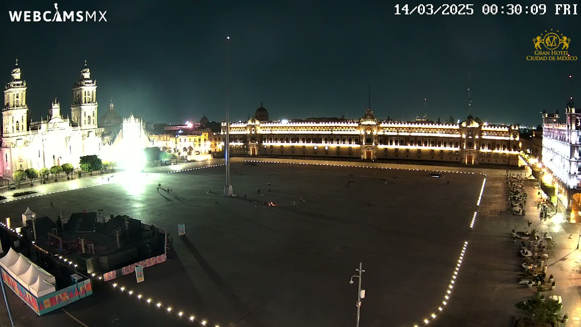 A large, illuminated plaza at night, featuring a cathedral and government buildings, with a few people sparsely scattered throughout.