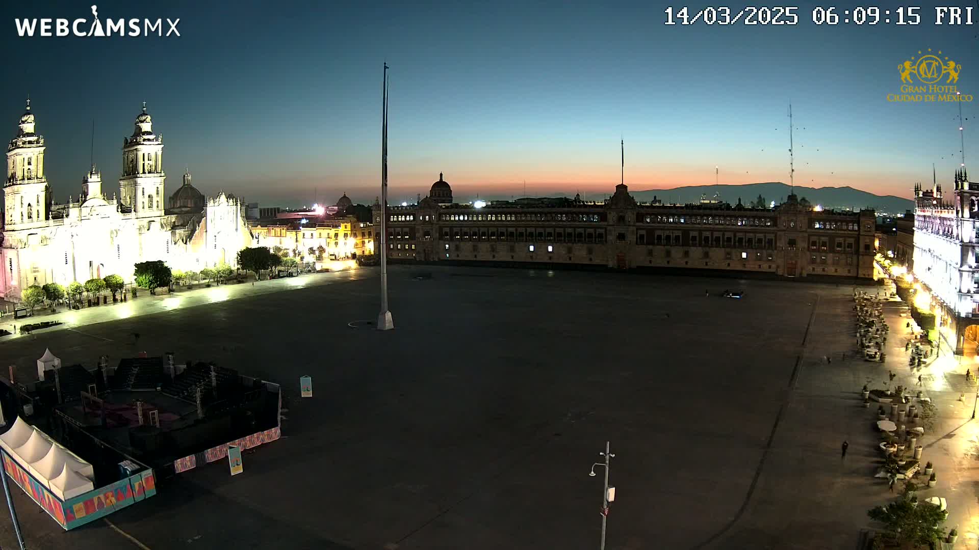 A large, mostly empty plaza at twilight is flanked by a grand cathedral and a long, rectangular building, with mountains visible in the distance under a clear, dusky sky.