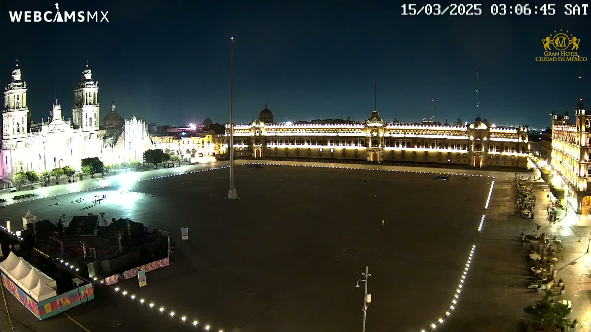 A large, mostly empty plaza at night is illuminated, with a cathedral and government building visible in the background under a clear, dark sky.