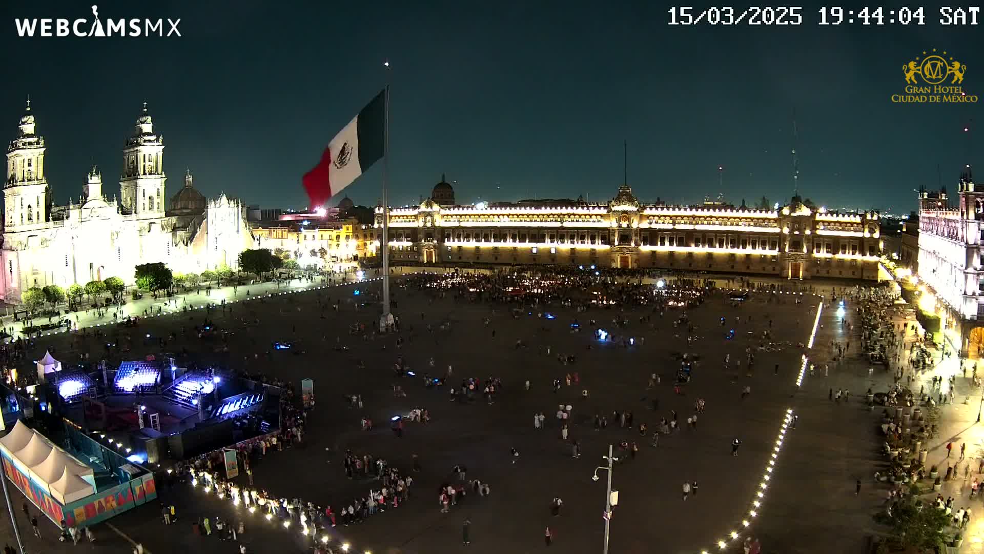 A brightly lit plaza at night, with a large Mexican flag, a cathedral, and government buildings, is filled with people.