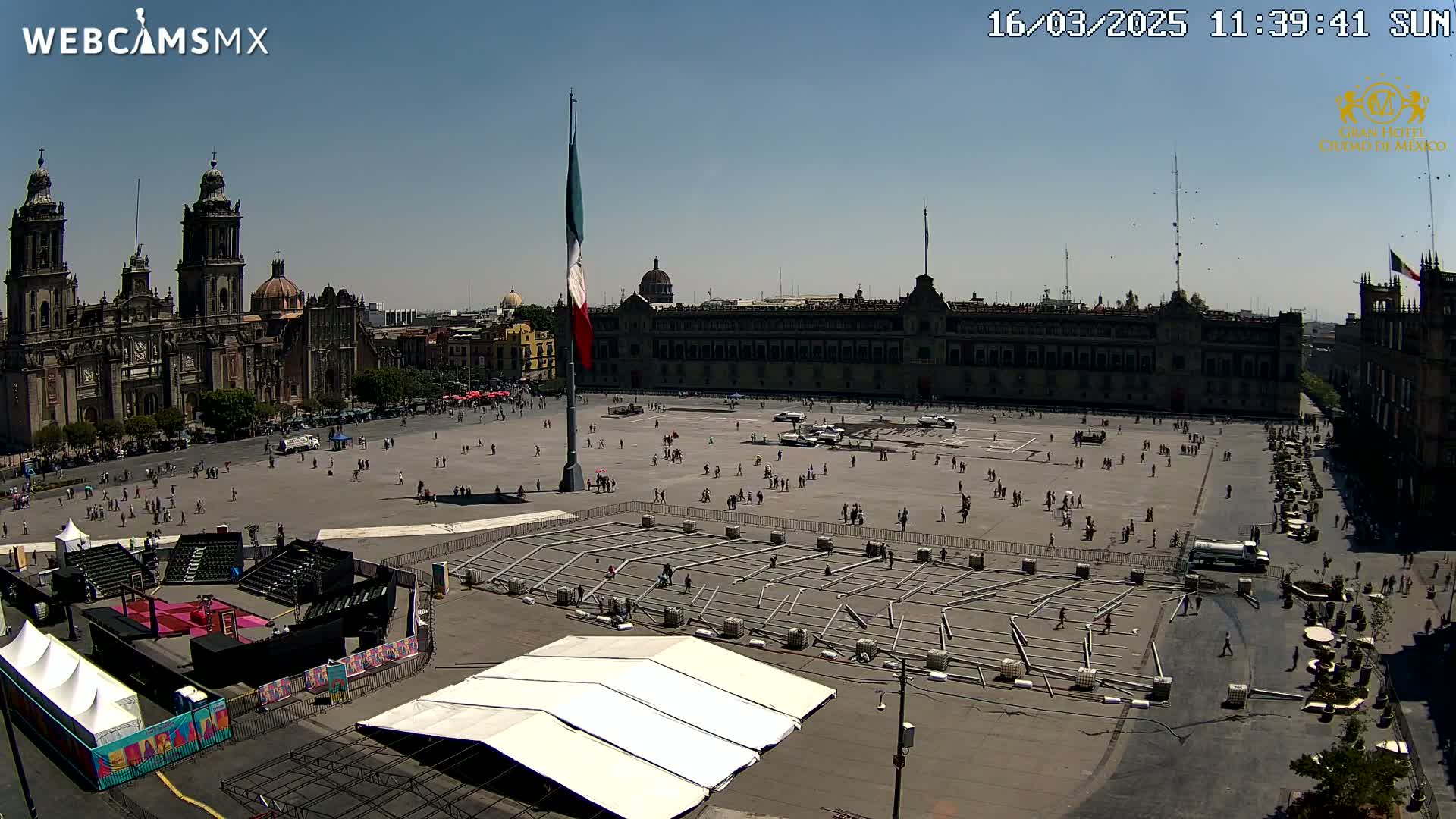 A large plaza with a cathedral and government building is seen under sunny skies; the plaza contains several temporary structures and a few people are present.