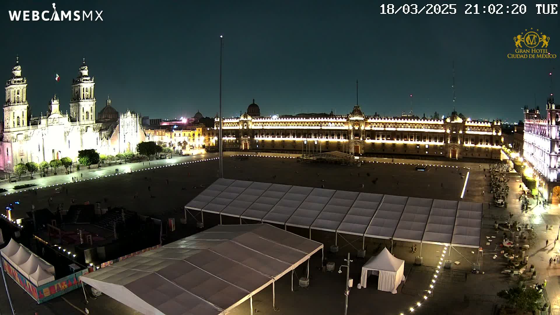 A nighttime, illuminated view of a large plaza with a cathedral and government building, featuring several temporary tents in the foreground.
