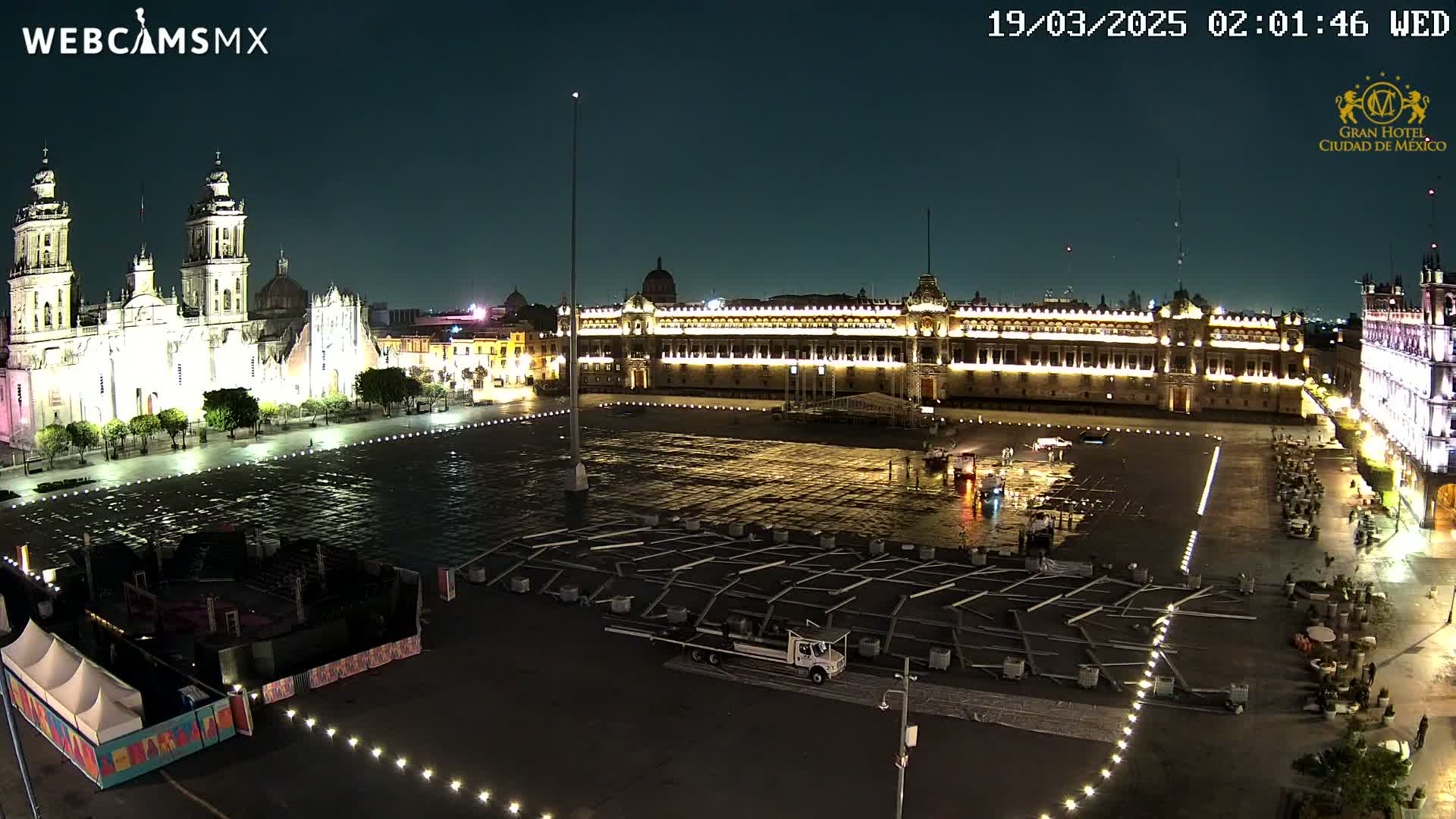 A large plaza at night, illuminated, showing a cathedral and government building, with a wet surface and some temporary structures.