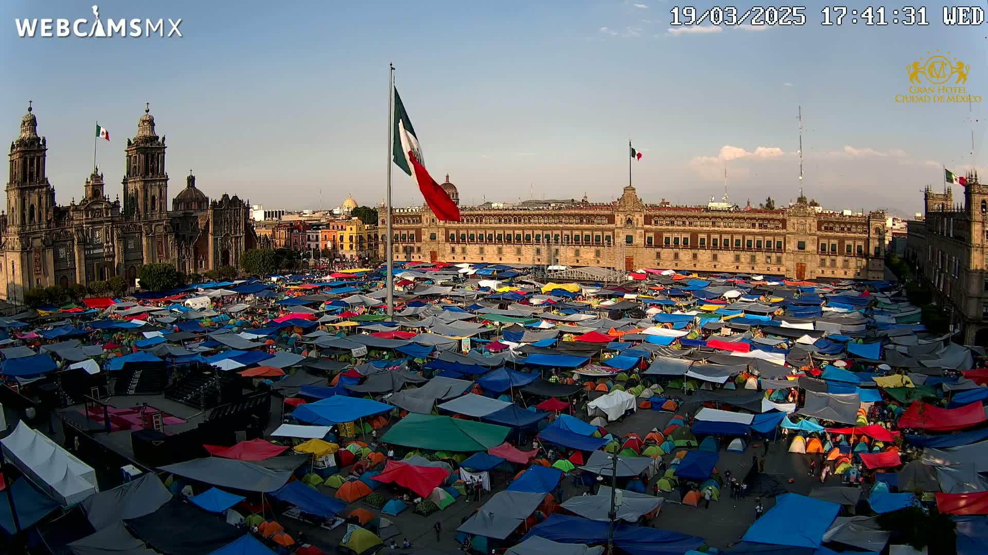 A large plaza is filled with numerous colorful tents under a mostly clear, late afternoon sky, with a cathedral and government building visible in the background.
