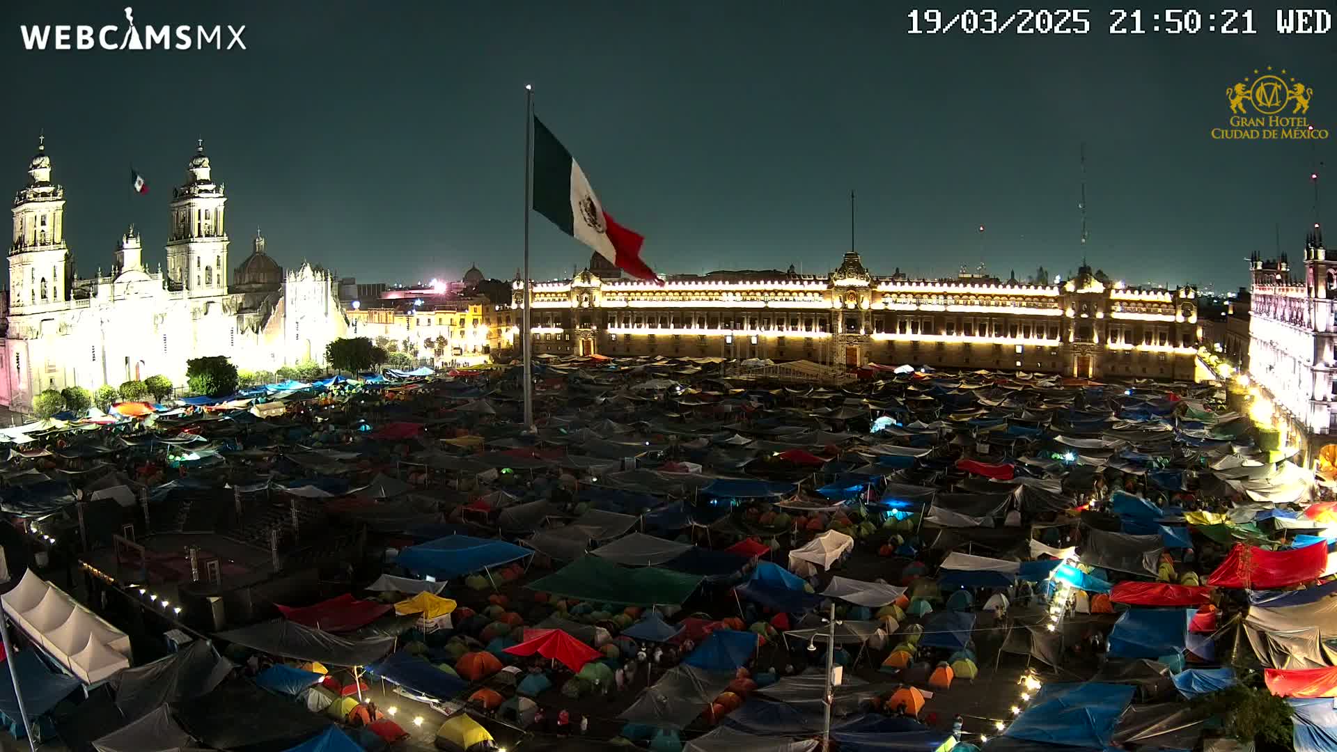 At night, a large plaza is filled with numerous tents, illuminated by lights, with a cathedral and a large building lit up in the background under a clear, dark sky.
