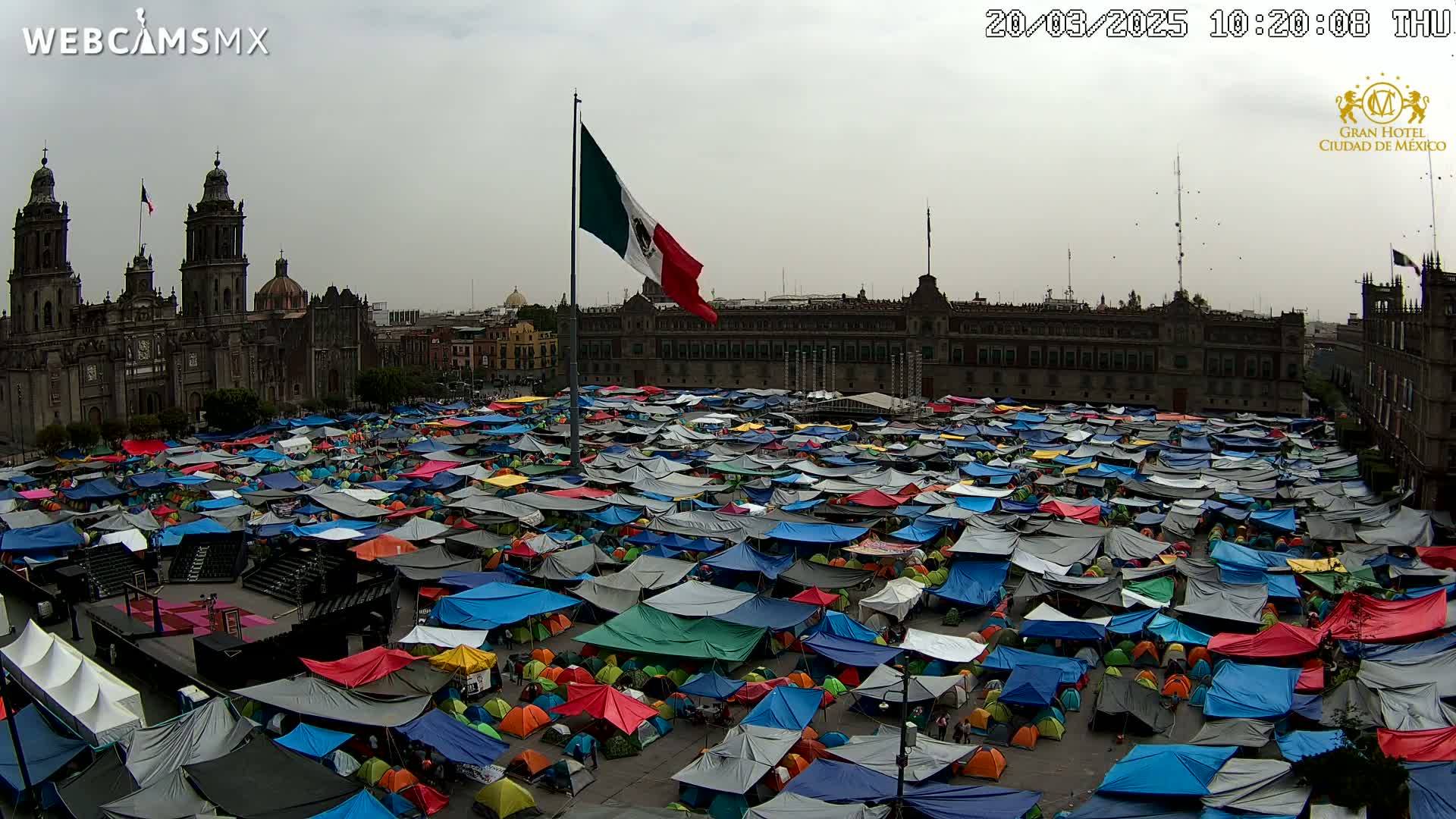 A large plaza is filled with numerous tents of various colors under an overcast sky, with a cathedral and government building in the background.