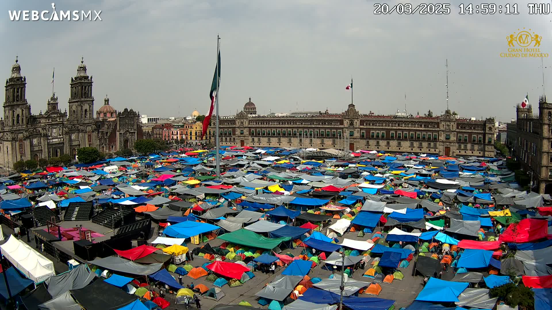 A large plaza filled with numerous colorful tents under a mostly cloudy sky, with a large building and cathedral visible in the background.