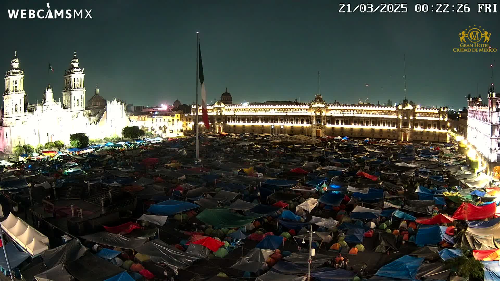 At night, a large plaza is filled with numerous tents surrounding illuminated historical buildings under a clear, dark sky.