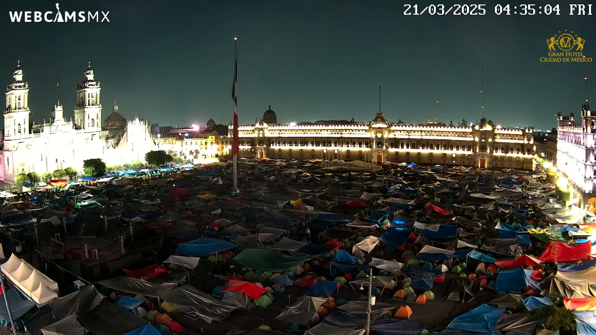 A large plaza at night, illuminated by lights on surrounding buildings, is filled with numerous tents.