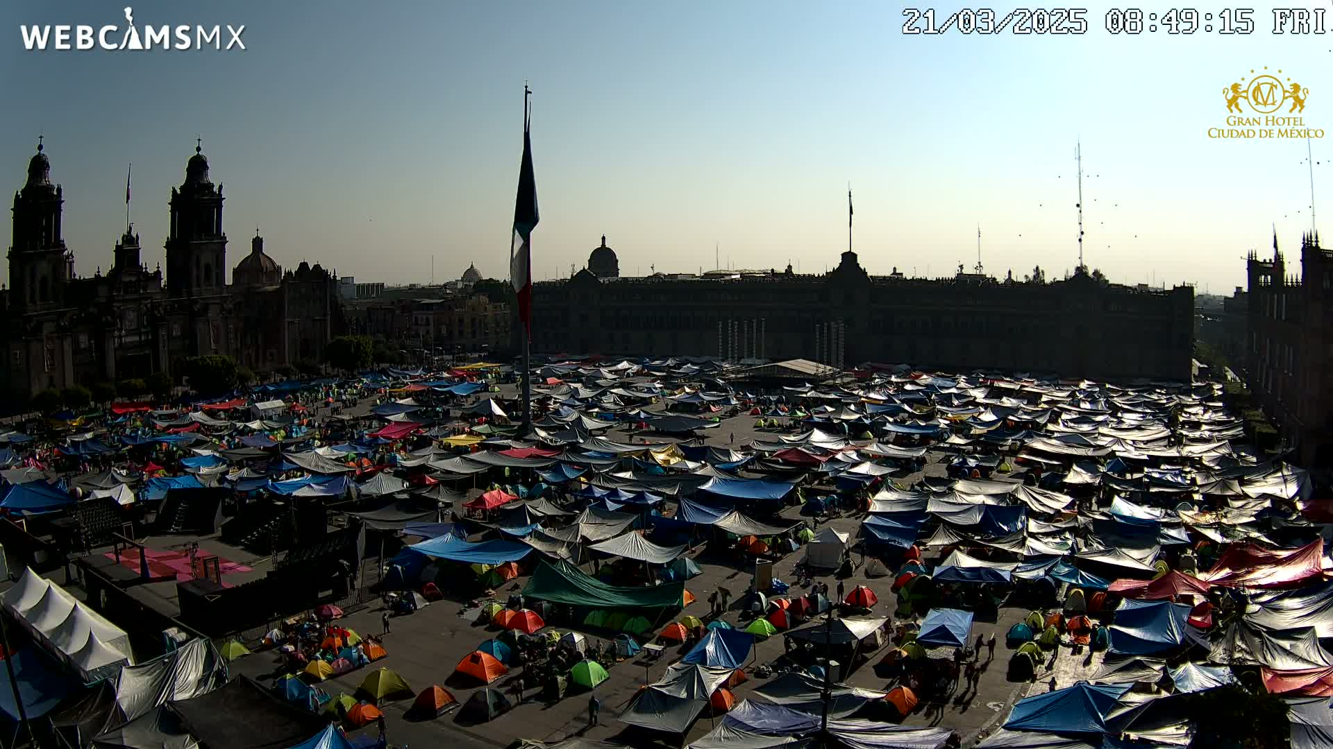 A large plaza is filled with numerous tents under a clear, sunny sky, with a large building and cathedral in the background.