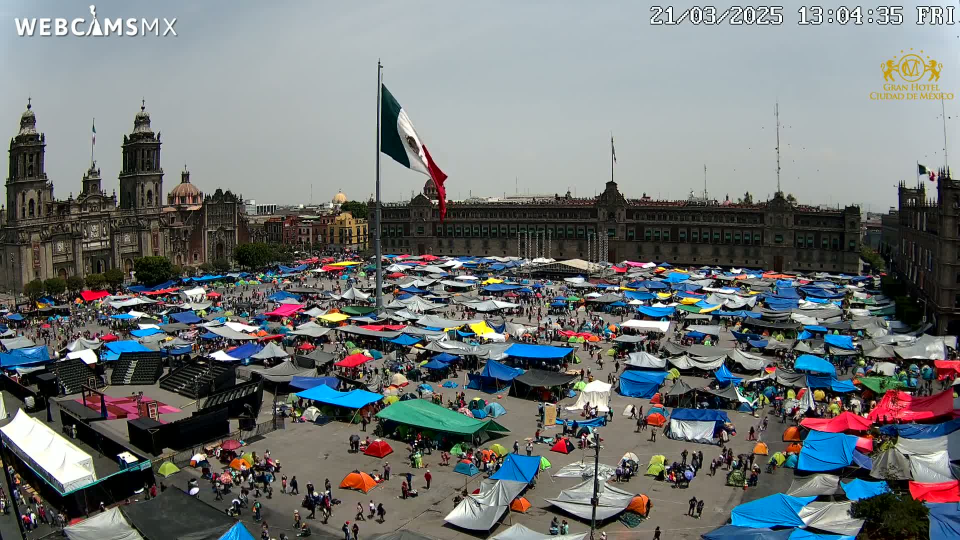 A large plaza, under a mostly clear sky, is filled with a dense collection of colorful tents and people in front of a historic building.