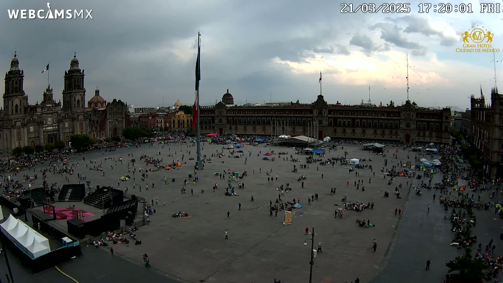 A large plaza, filled with many people and some tents, is situated in front of a grand cathedral and palace under an overcast sky.