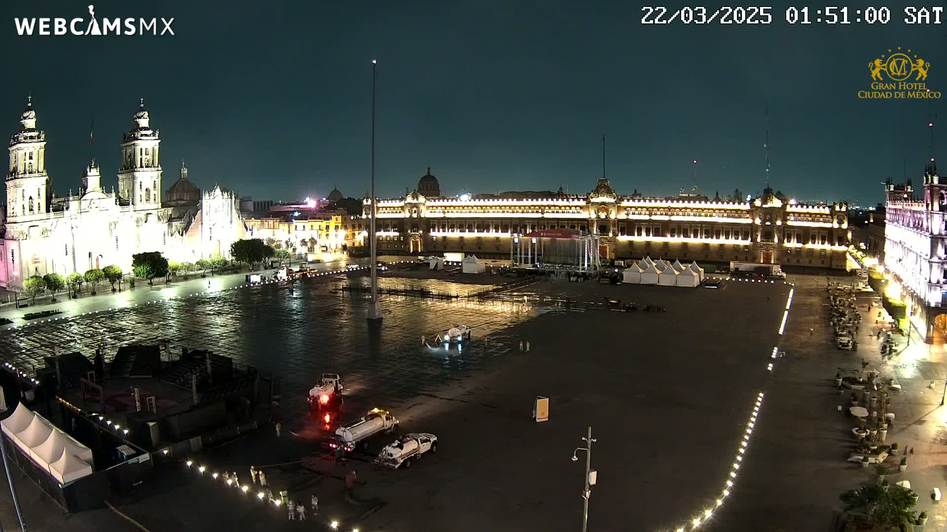 A large, mostly empty plaza at night, illuminated by lights on surrounding buildings, including a cathedral and government buildings, is being cleaned by several trucks.
