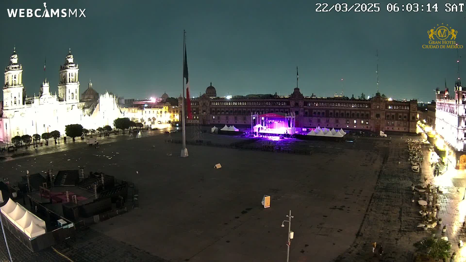A large, mostly empty plaza at night is lit by the surrounding buildings, including a brightly lit cathedral and palace, with a stage set up in the center and a Mexican flagpole standing tall.