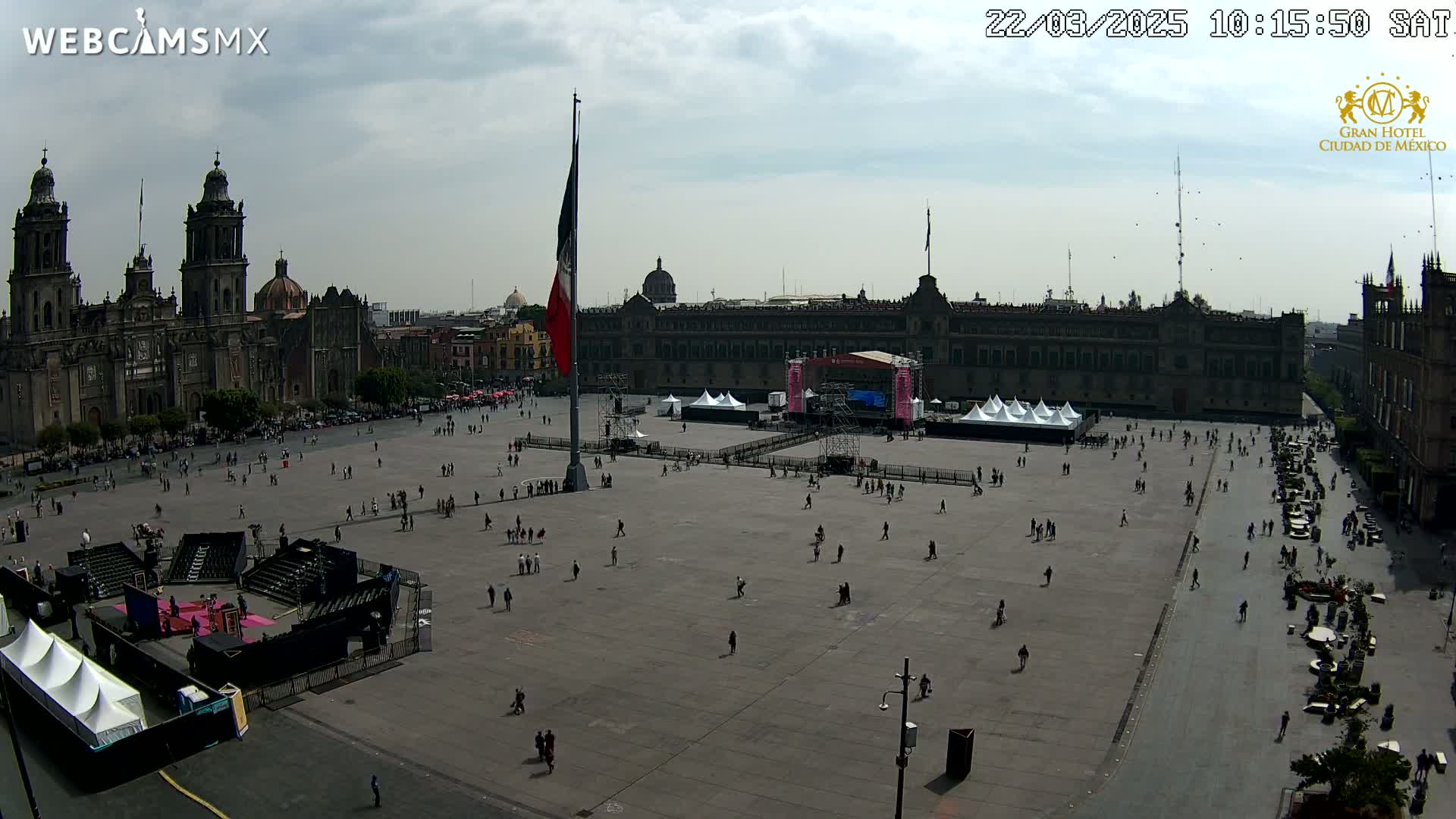 A large plaza with a few people scattered throughout, a stage setup, a Mexican flag on a pole, and a cathedral in the background under a mostly cloudy sky.
