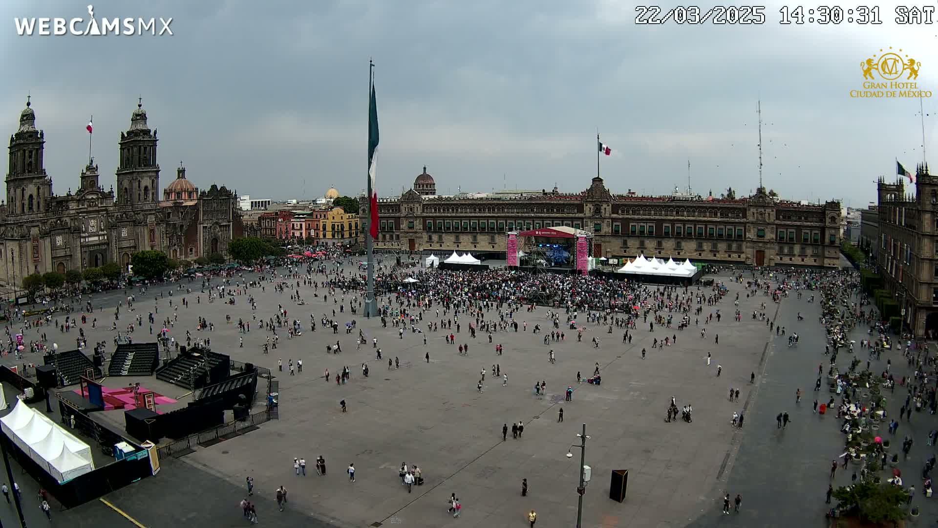 A large plaza with a crowd of people gathered around a stage, situated between a cathedral and a government building under an overcast sky.