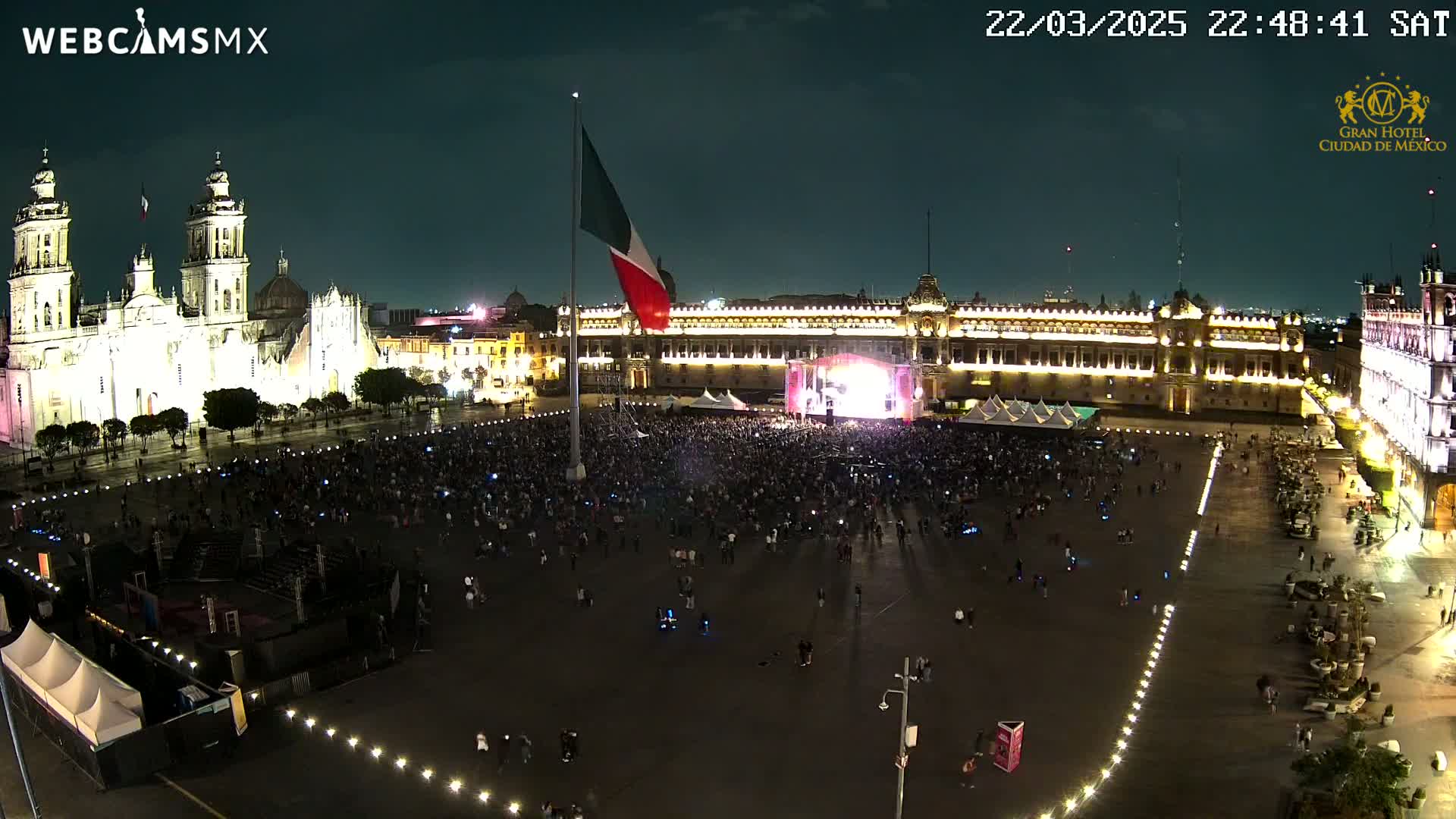 A large crowd gathers in a brightly lit plaza at night, in front of a stage and buildings, with a large Mexican flag on a flagpole in the center.