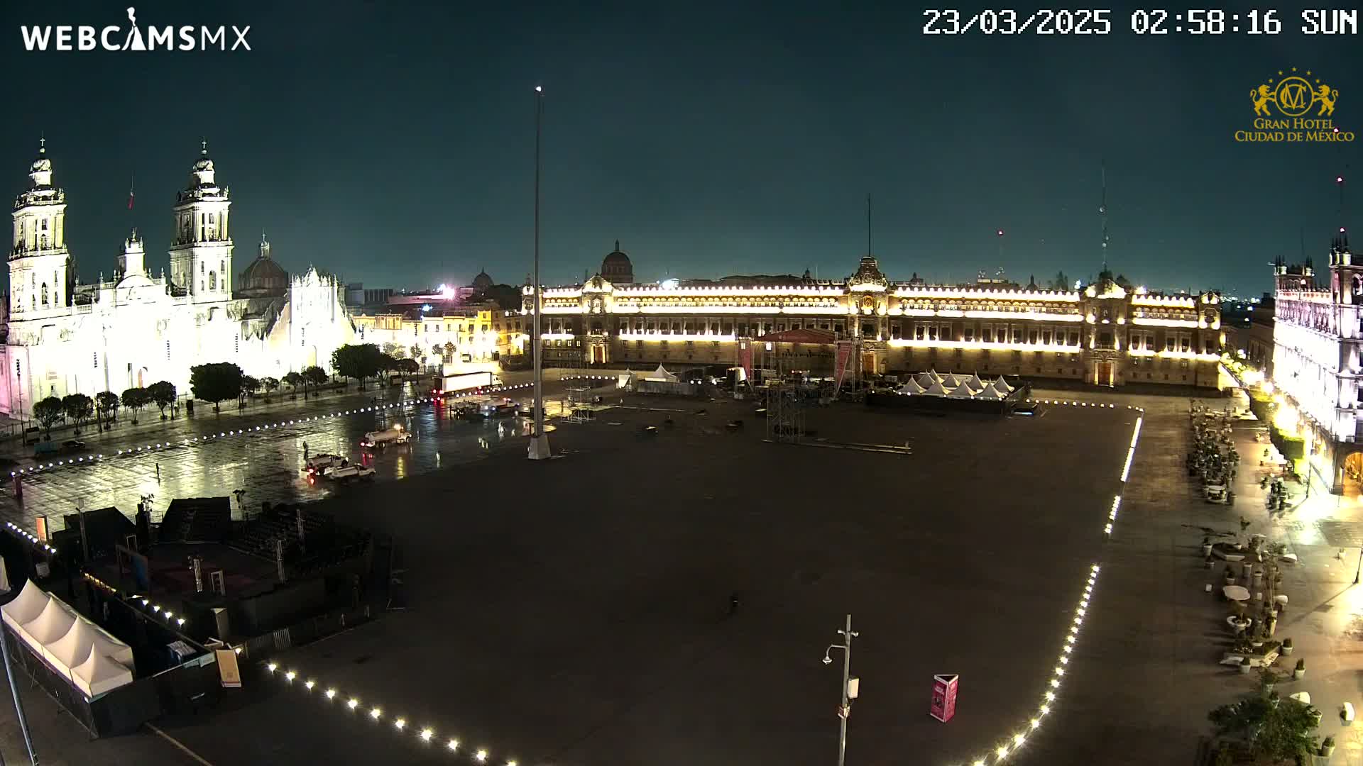 A nighttime, wet view of a large plaza with a cathedral and government buildings illuminated, showing some vehicles and sparse pedestrian traffic.