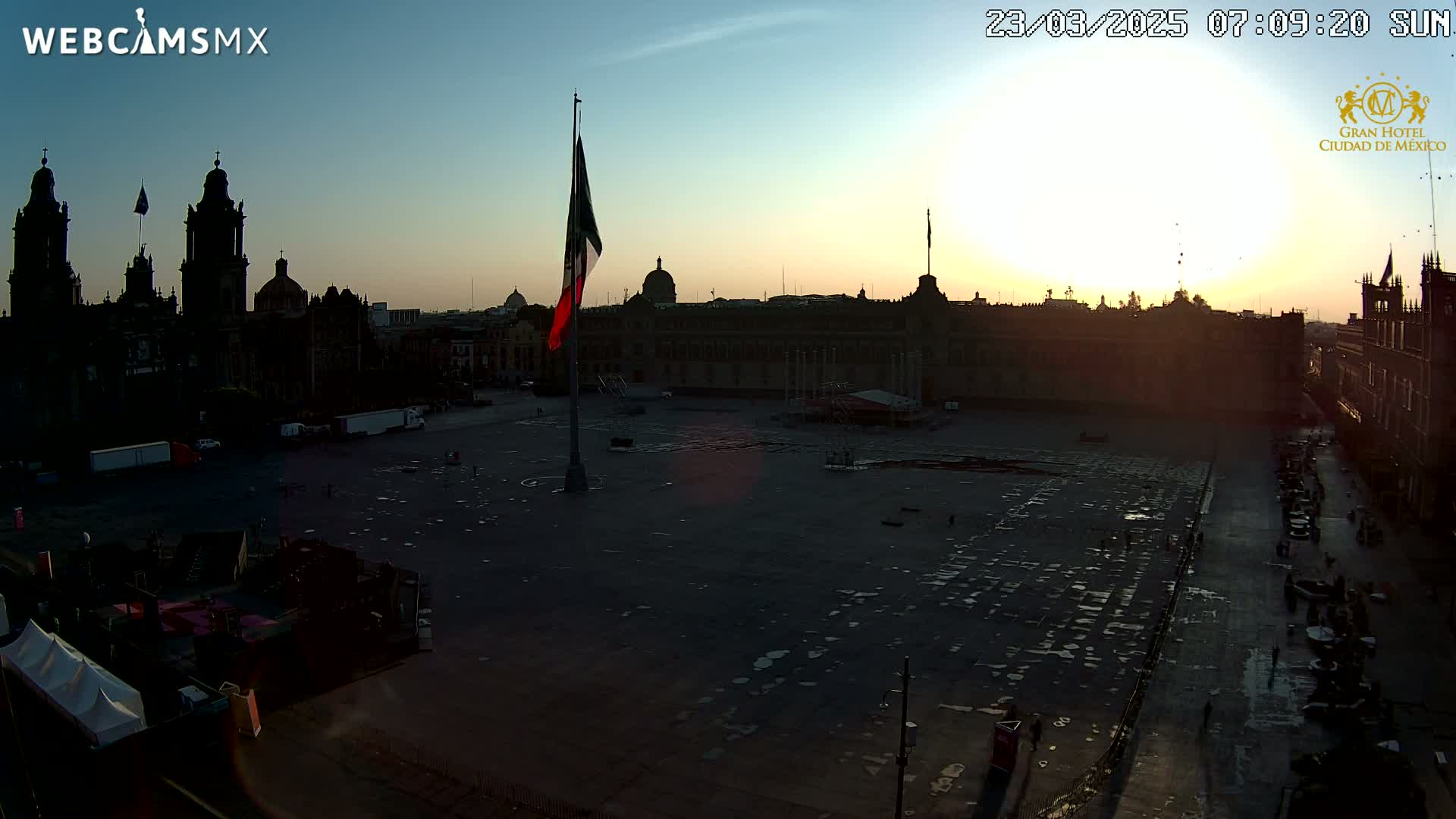 A large, mostly empty plaza at sunrise, with a Mexican flag raised high, is surrounded by historical buildings including a cathedral.