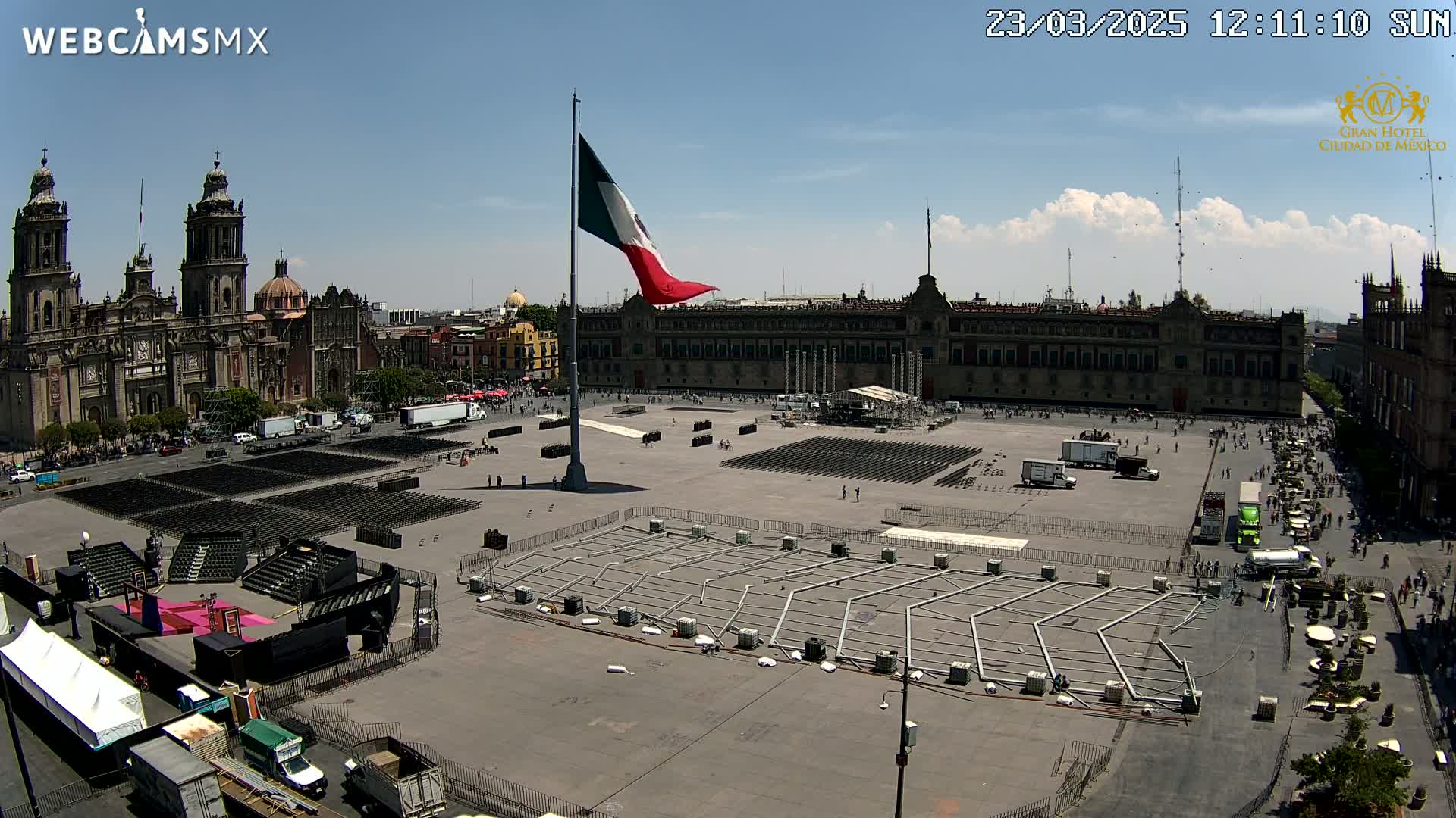 A large plaza is being prepared for an event, with a stage being set up, rows of chairs arranged, and the Mexican flag flying high on a flagpole in front of a large building under a mostly sunny sky.