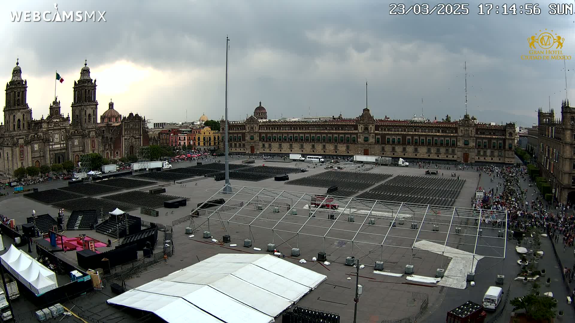 A large plaza is being set up for an event, under a cloudy sky, featuring numerous rows of empty chairs and large tents in front of a cathedral and a large government building.