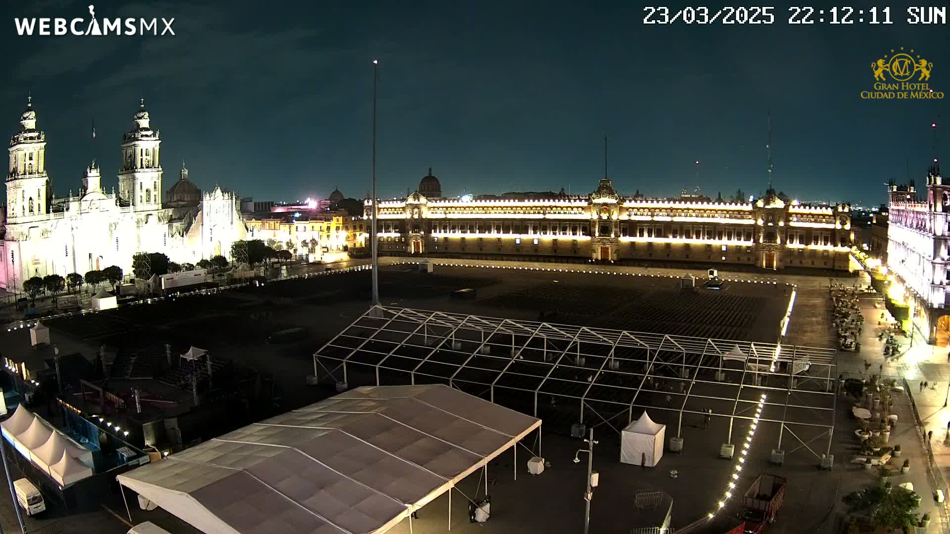 A large, illuminated plaza at night features a cathedral and government building, with several temporary structures set up in the square.