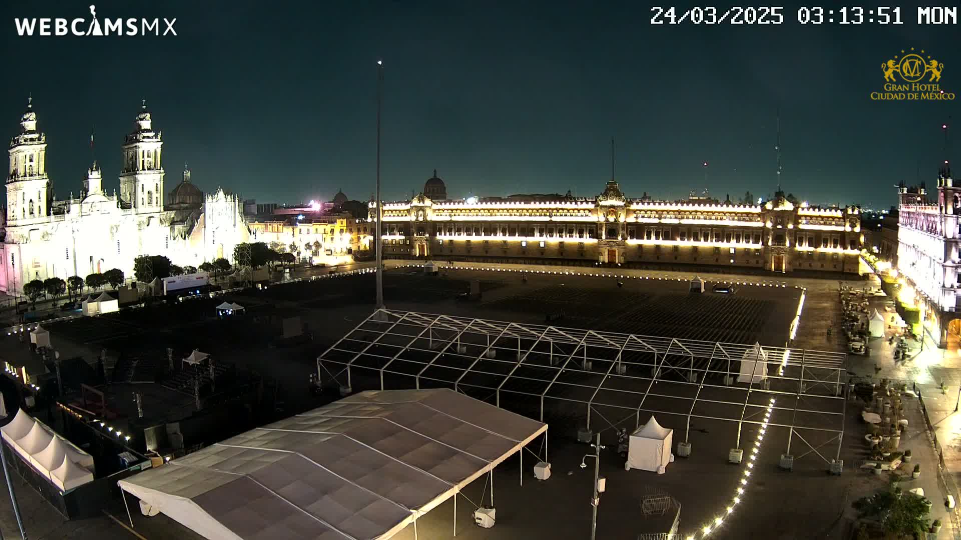 A nighttime, high-angle view of a large plaza with several illuminated buildings, including a cathedral and a palace, with numerous temporary structures set up in the square.