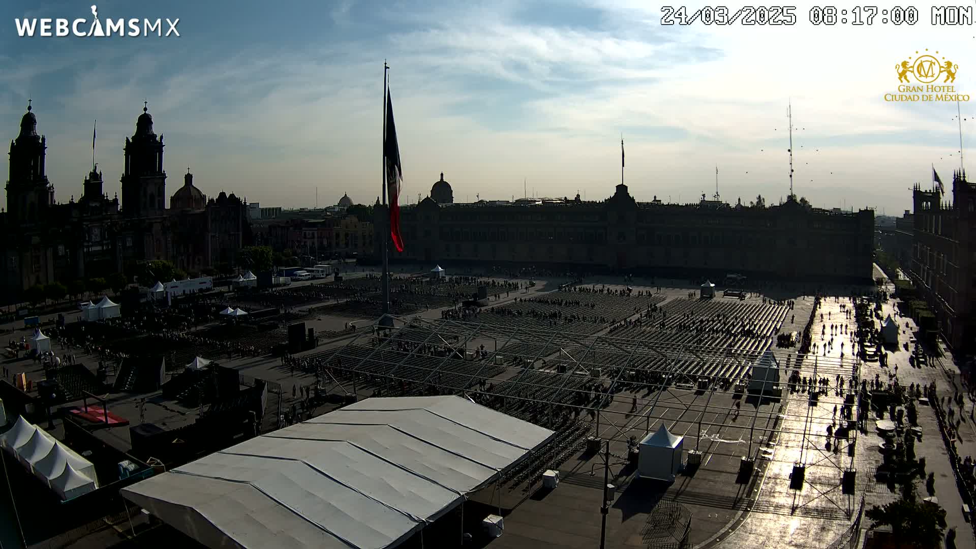 A large plaza with numerous rows of chairs set up under a mostly sunny sky, surrounded by large, silhouetted buildings.