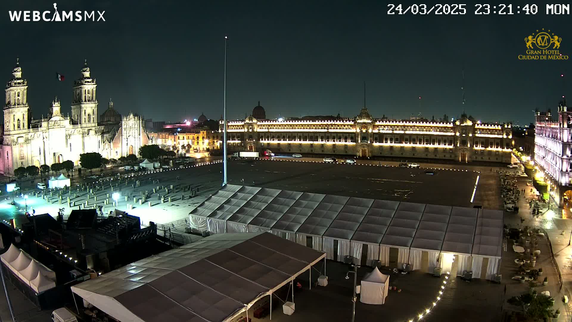 A large, empty plaza at night is surrounded by illuminated buildings, including a cathedral and a palace, with numerous white tents set up in the foreground.