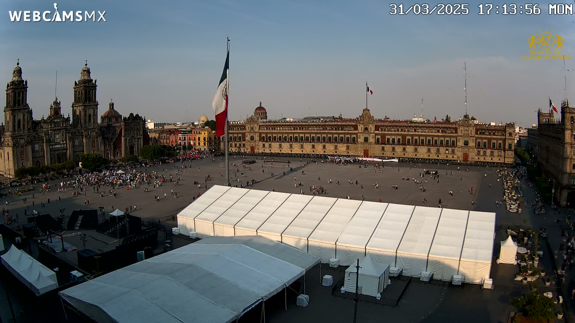A large plaza with a cathedral and government building in the background, under a mostly clear sky, contains numerous people and several large white tents.