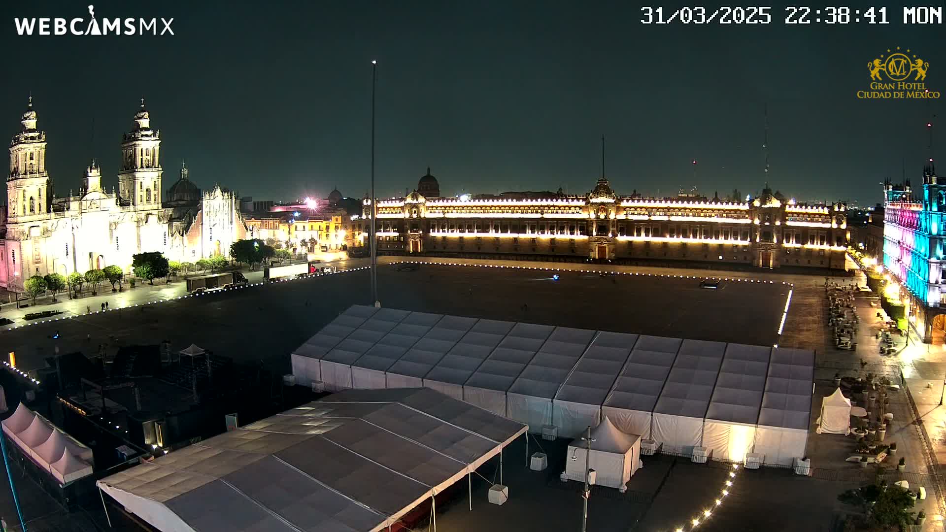 A nighttime view of a large plaza with a cathedral and government building illuminated, featuring several tents set up in the foreground under a clear sky.