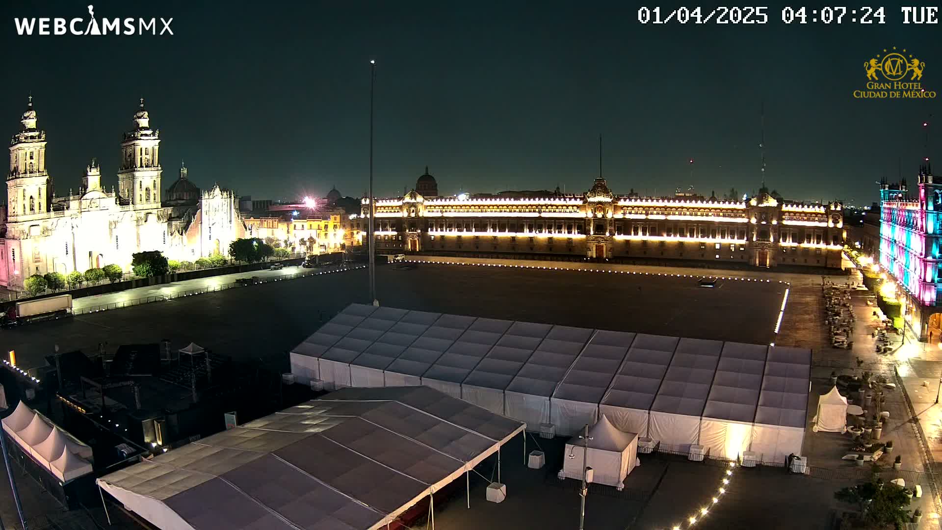 At night, a large plaza contains several large white tents and is bordered by illuminated historical buildings.