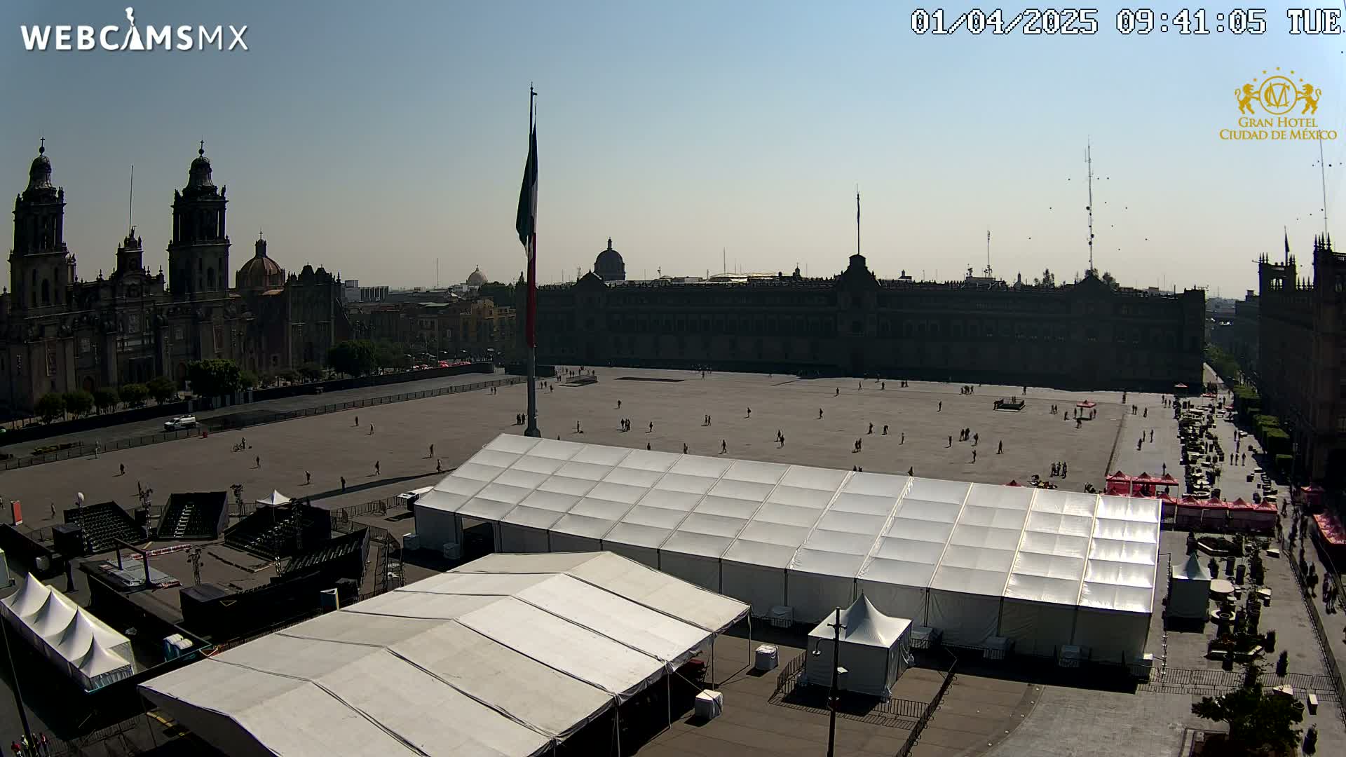 A large plaza with several white tents and a Mexican flagpole, overlooked by a cathedral and a large building, under a clear sunny sky.