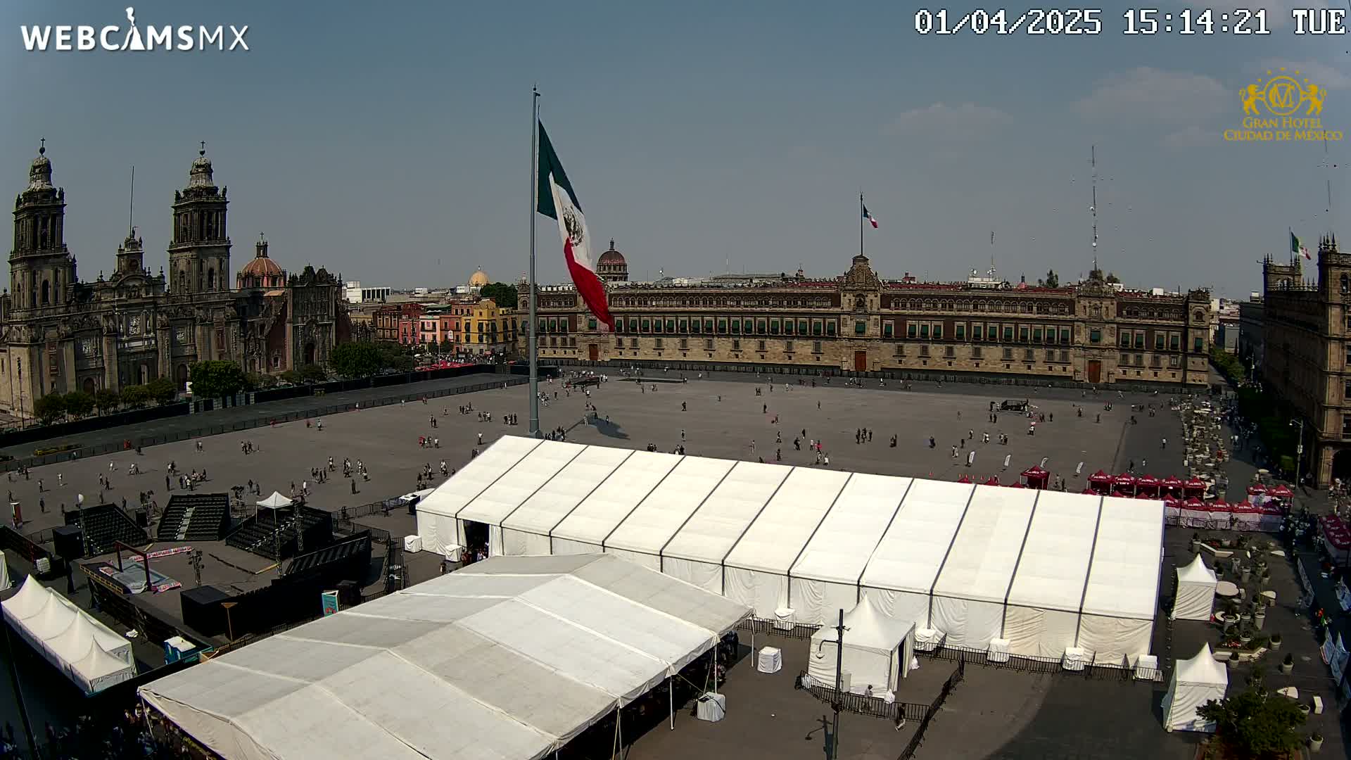A large plaza with several white tents, a cathedral, and a government building is visible under a sunny sky.