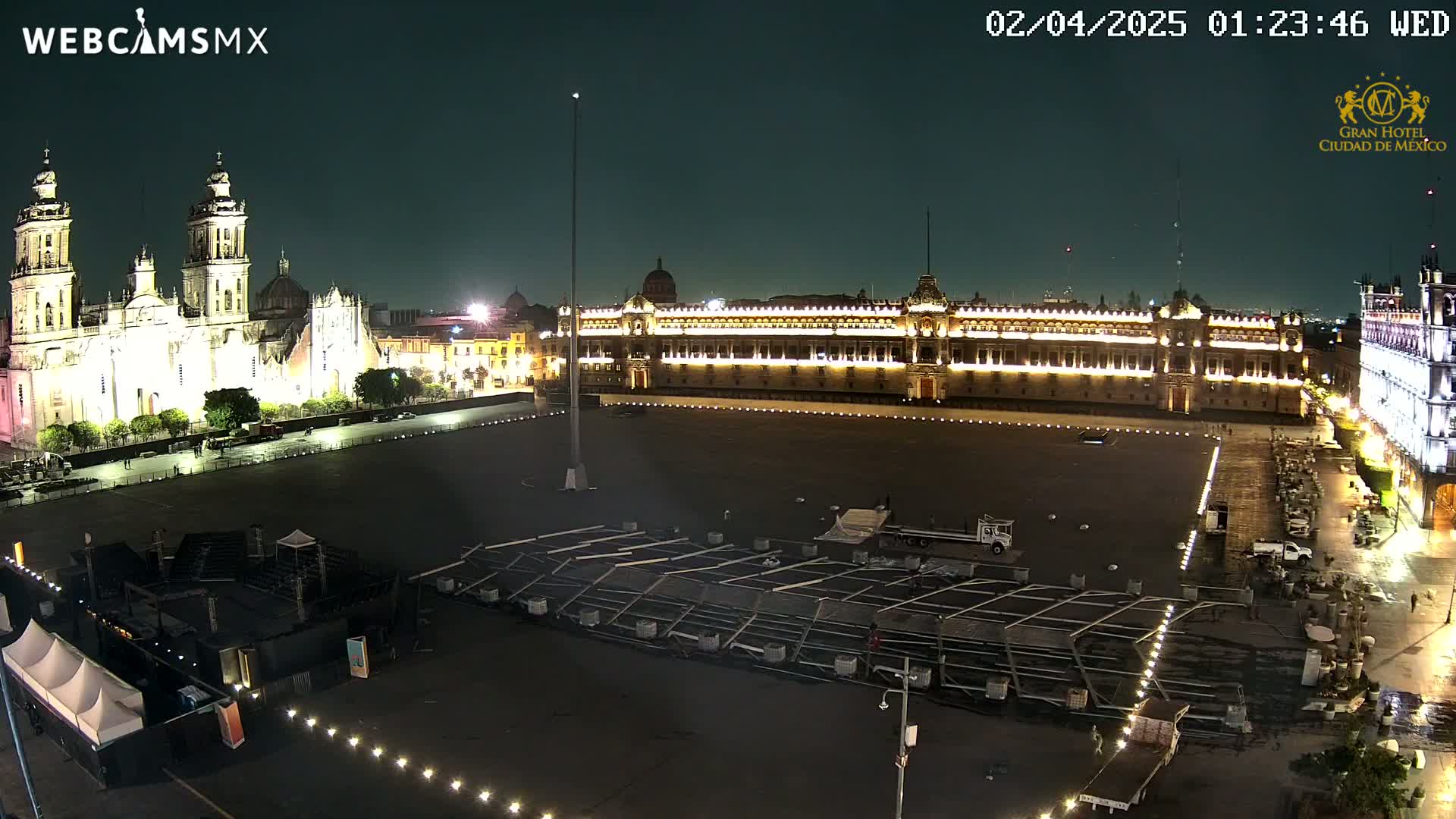 A large, empty plaza at night is illuminated, showing a cathedral and a long, brightly lit building.