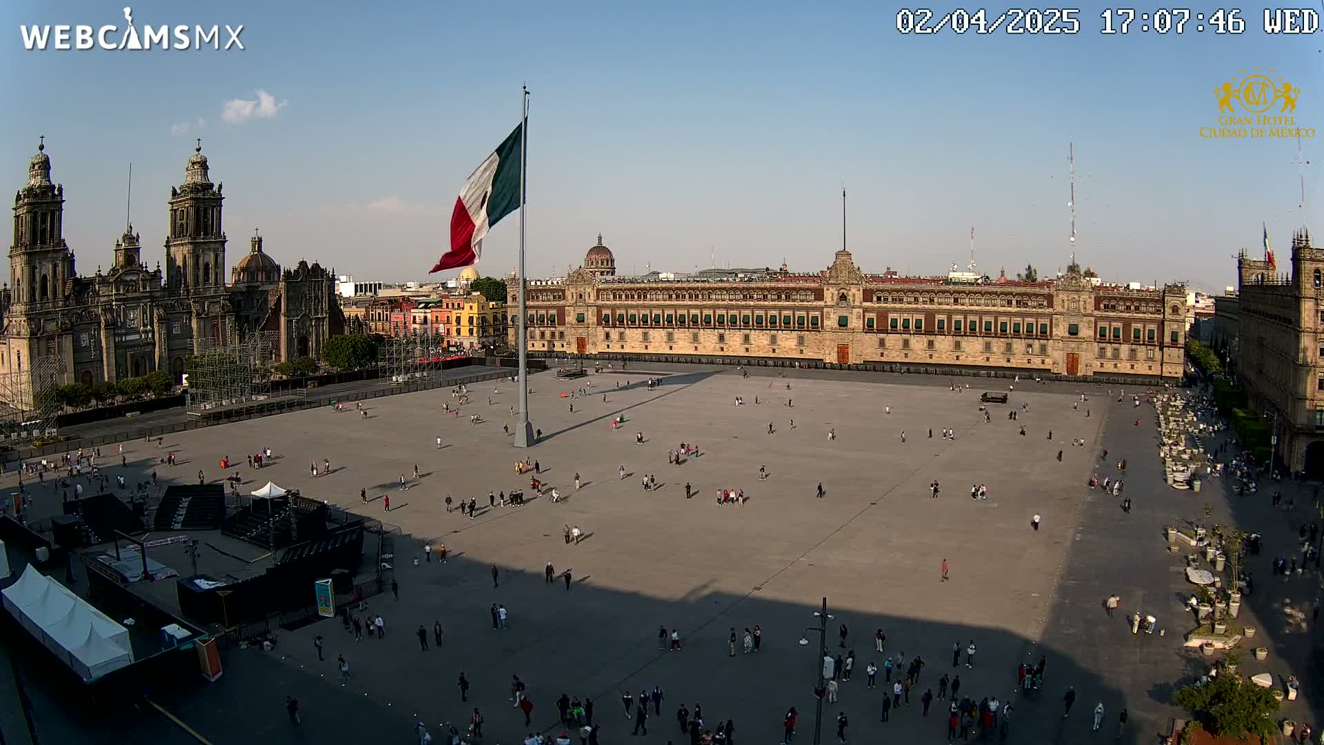 A large plaza in Mexico City, under a clear sky, is sparsely populated with people, featuring the Metropolitan Cathedral and National Palace in the background, with the Mexican flag flying high.