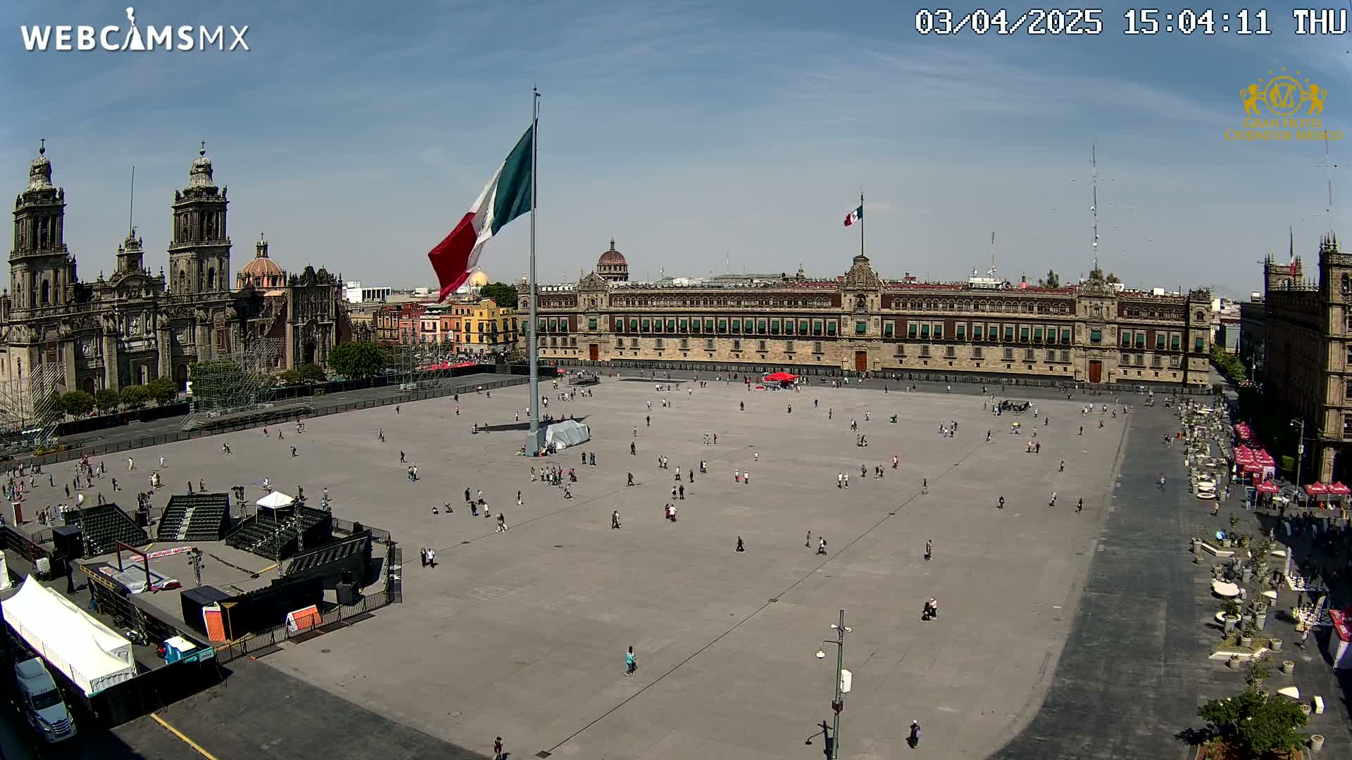 A large plaza, mostly empty except for scattered people, is situated between a cathedral and a long government building under a sunny sky.