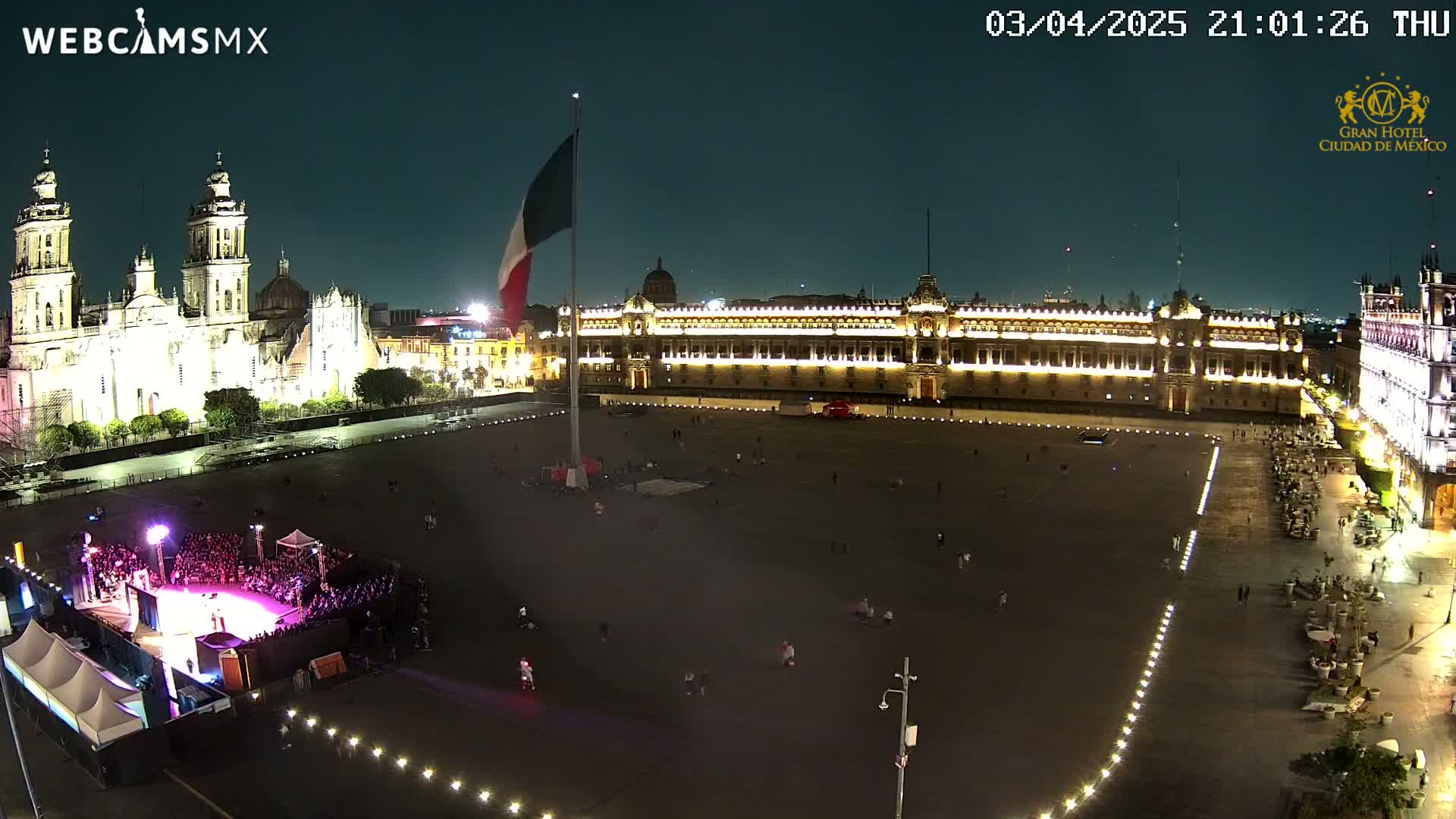 A large, mostly empty plaza at night is illuminated, with a cathedral and government building visible in the background under a clear, dark sky.