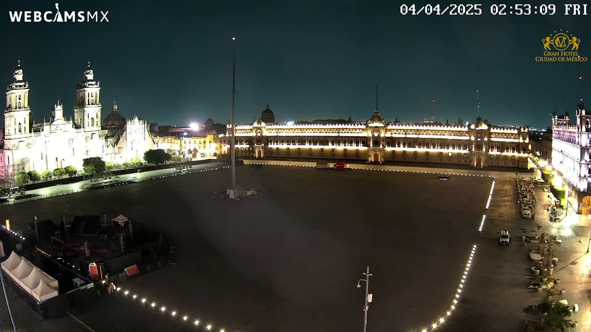 A large, mostly empty plaza at night, illuminated by lights, is flanked by a cathedral and a large, ornate building.