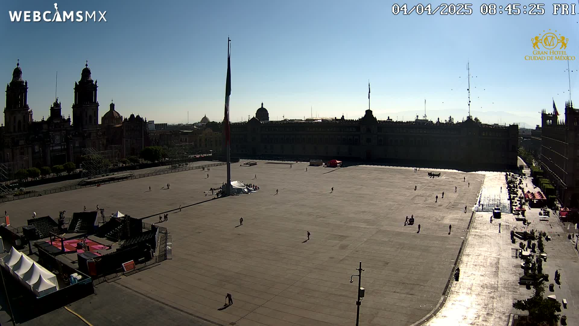 A large plaza with a flagpole and several buildings, including a cathedral, is mostly empty under a clear, sunny sky.