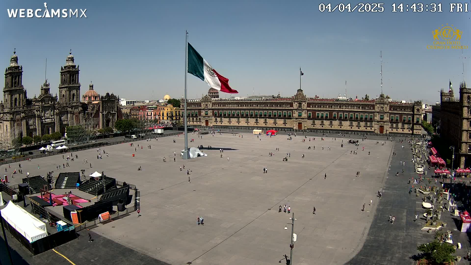 A large plaza with a few people, a Mexican flag on a tall pole, a cathedral, and a large government building under a clear, sunny sky.