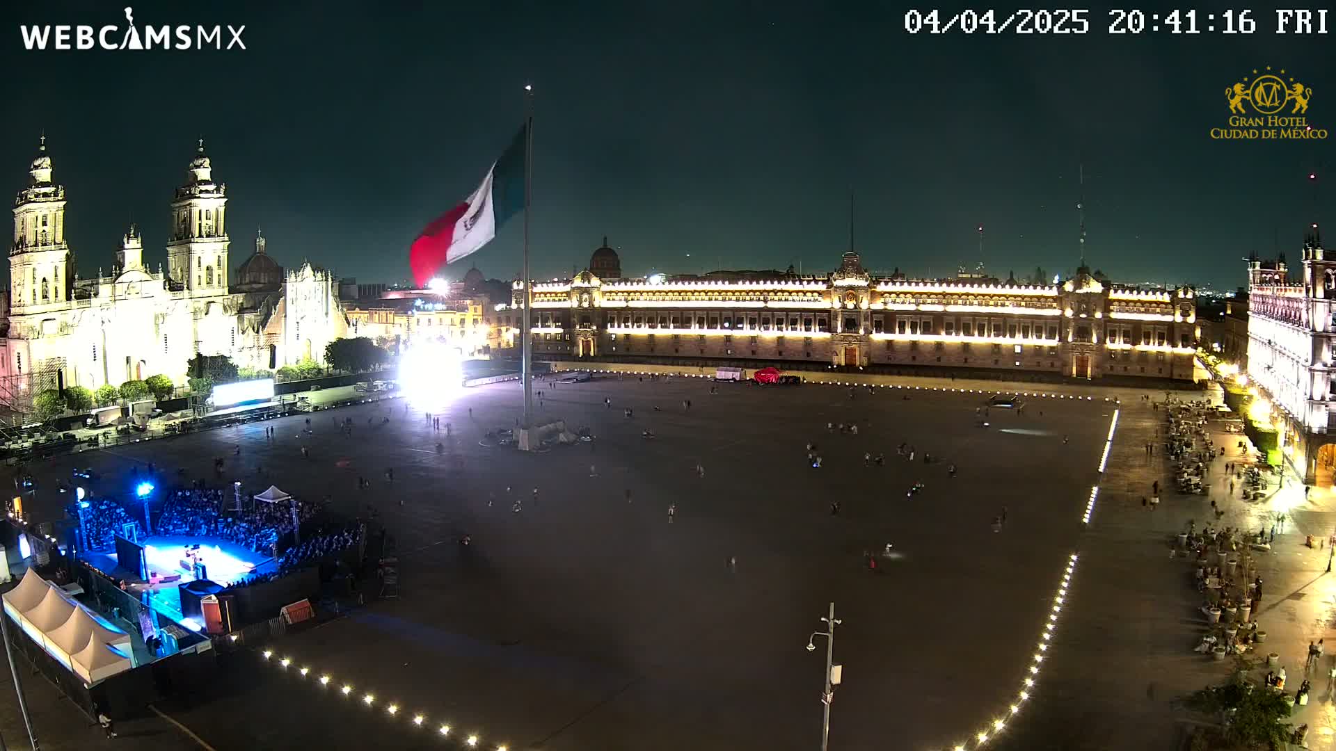 A large, illuminated plaza at night is shown, featuring a cathedral, government building, and a Mexican flag flying on a tall pole, with a small crowd gathered for an event.