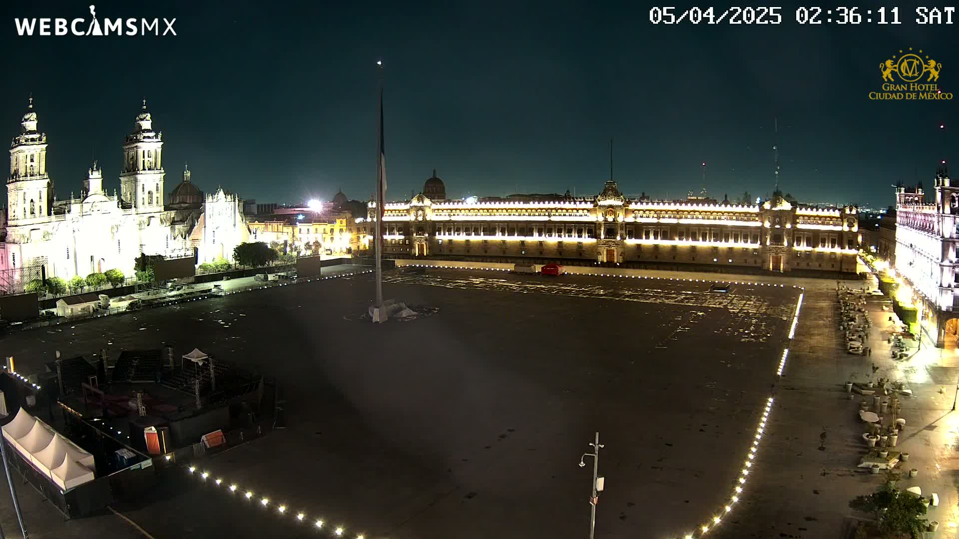A large, mostly empty plaza at night, illuminated by lights, is flanked by a grand cathedral and palace.