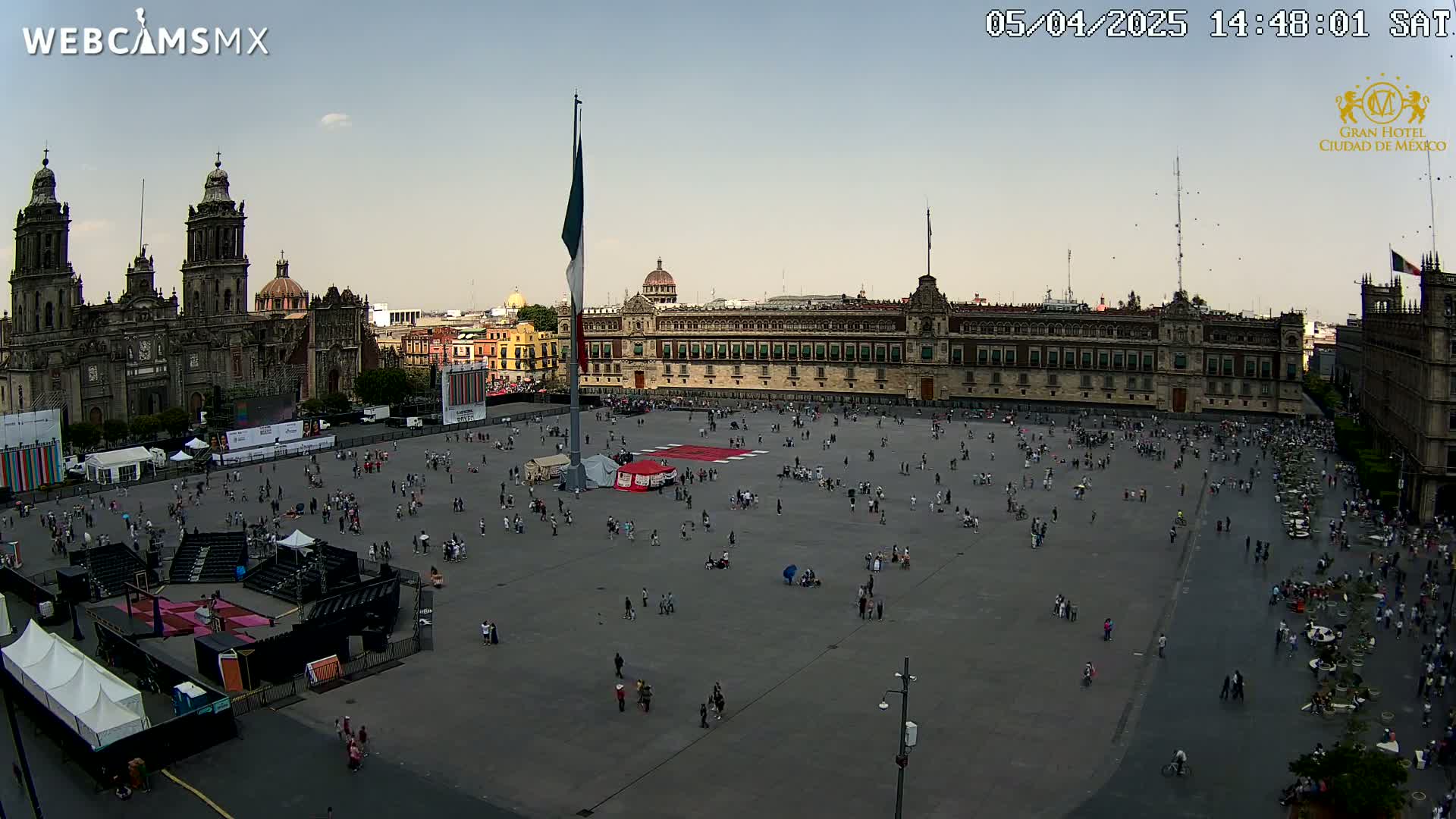 A large plaza with a sparsely scattered crowd, flanked by a cathedral and a long government building, under a mostly clear sky.