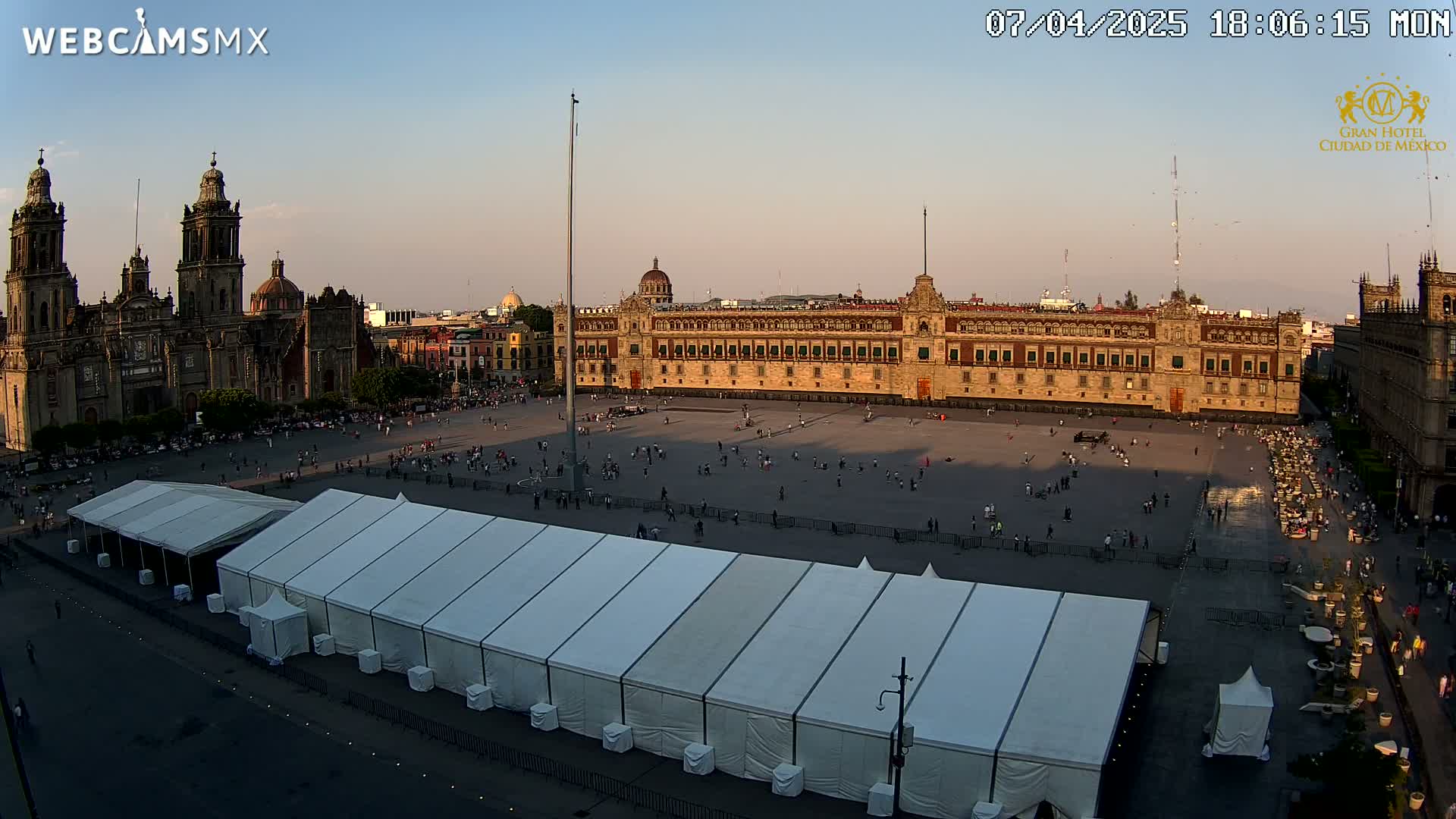 A large plaza with a long building, a cathedral, and several white tents is shown under a clear, dusky sky.
