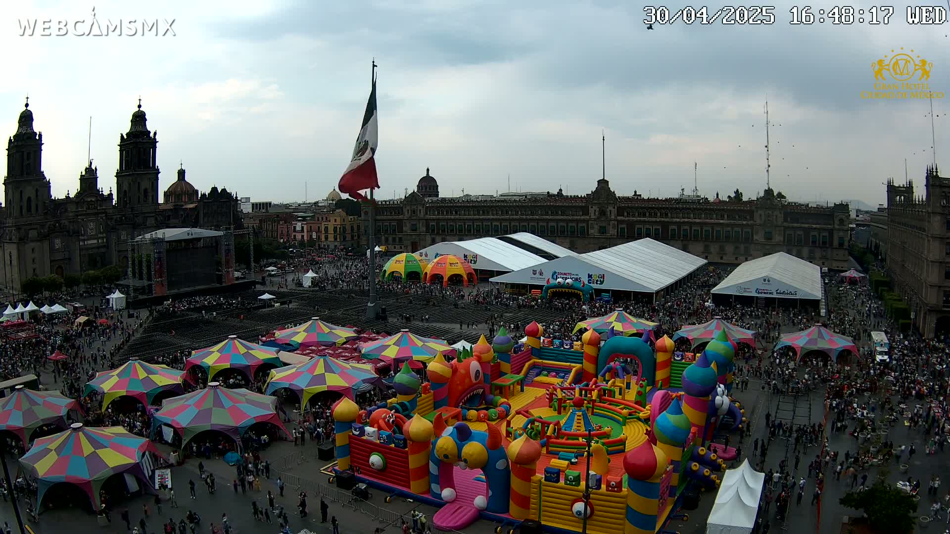 A large plaza is filled with people, numerous colorful tents and booths, and a giant inflatable playground, all under an overcast sky.
