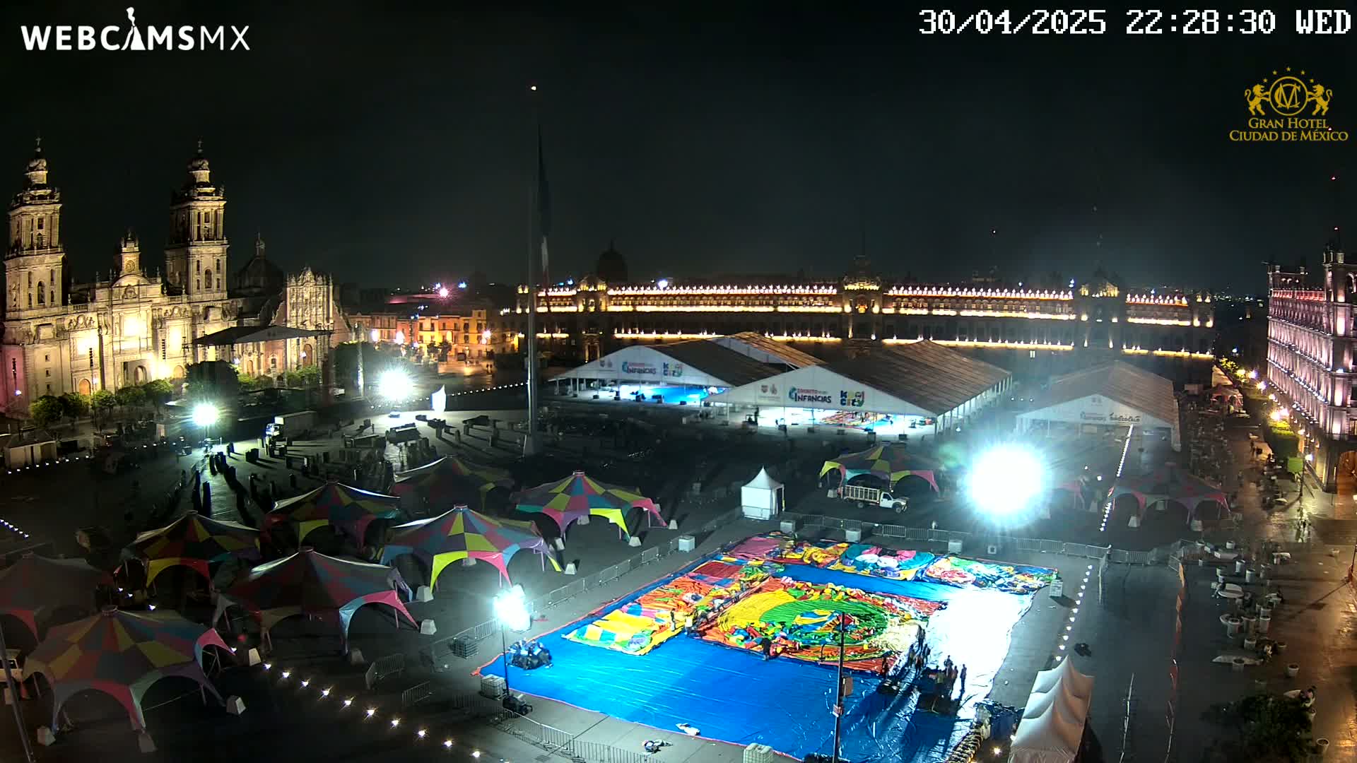 A nighttime aerial view of a large plaza with a cathedral, government buildings, temporary tents, and a large inflatable playground.