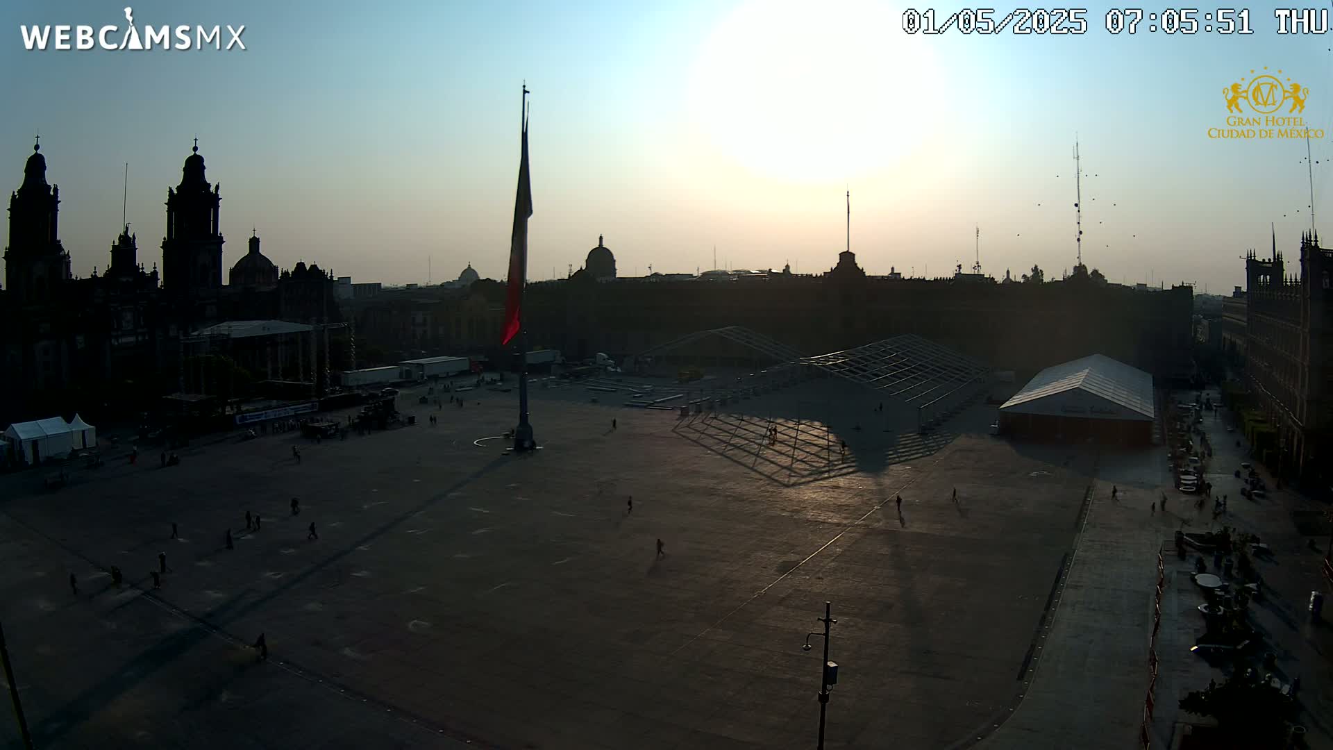 A large plaza with a few people and several buildings is silhouetted against a sunrise.