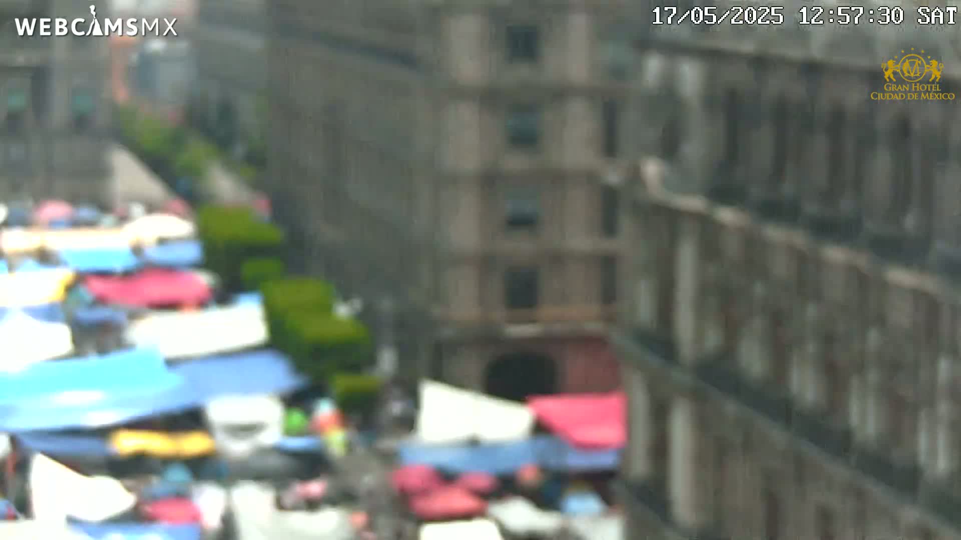 A blurry, high-angle view of a street market with colorful stalls and a tall building in the background.